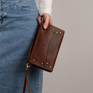 Peut inclure: Une pochette en cuir marron avec une sangle, orn&eacute;e de rivets et d'un &eacute;l&eacute;ment de design central. La pochette est tenue par une personne portant un haut blanc &agrave; manches longues et un jean bleu.
