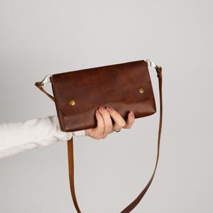 Peut inclure: Un sac bandouli&egrave;re en cuir marron avec une longue bandouli&egrave;re r&eacute;glable. Le sac a une fermeture &agrave; rabat s&eacute;curis&eacute;e par deux boutons-pression couleur laiton. Le sac est tenu par une personne sur un fond uni.