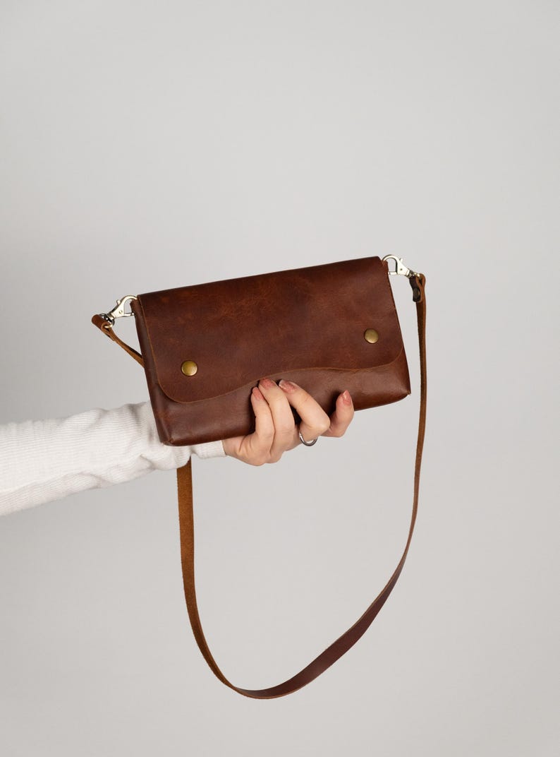 Peut inclure: Un sac bandouli&egrave;re en cuir marron avec une longue bandouli&egrave;re r&eacute;glable. Le sac a une fermeture &agrave; rabat s&eacute;curis&eacute;e par deux boutons-pression couleur laiton. Le sac est tenu par une personne sur un fond uni.