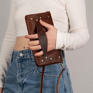Peut inclure: Un portefeuille en cuir marron avec une poign&eacute;e et une bandouli&egrave;re. Le portefeuille rectangulaire est fix&eacute; avec des rivets. La poign&eacute;e est fix&eacute;e avec des rivets. La bandouli&egrave;re est une fine corde en cuir marron.