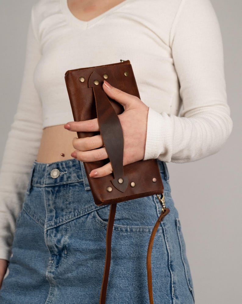 Peut inclure: Un portefeuille en cuir marron avec une poign&eacute;e et une bandouli&egrave;re. Le portefeuille rectangulaire est fix&eacute; avec des rivets. La poign&eacute;e est fix&eacute;e avec des rivets. La bandouli&egrave;re est une fine corde en cuir marron.