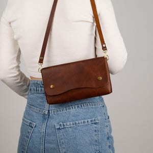 Peut inclure: Un sac bandouli&egrave;re en cuir marron avec une fermeture &agrave; rabat et deux boutons dor&eacute;s. Le sac est dot&eacute; d'une longue bandouli&egrave;re r&eacute;glable. Le sac est port&eacute; &agrave; l'&eacute;paule.