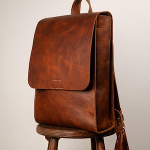 May include: A brown leather backpack with a flap closure, handle, and adjustable straps. The bag is sitting on a wooden stool. The leather has a textured appearance.
