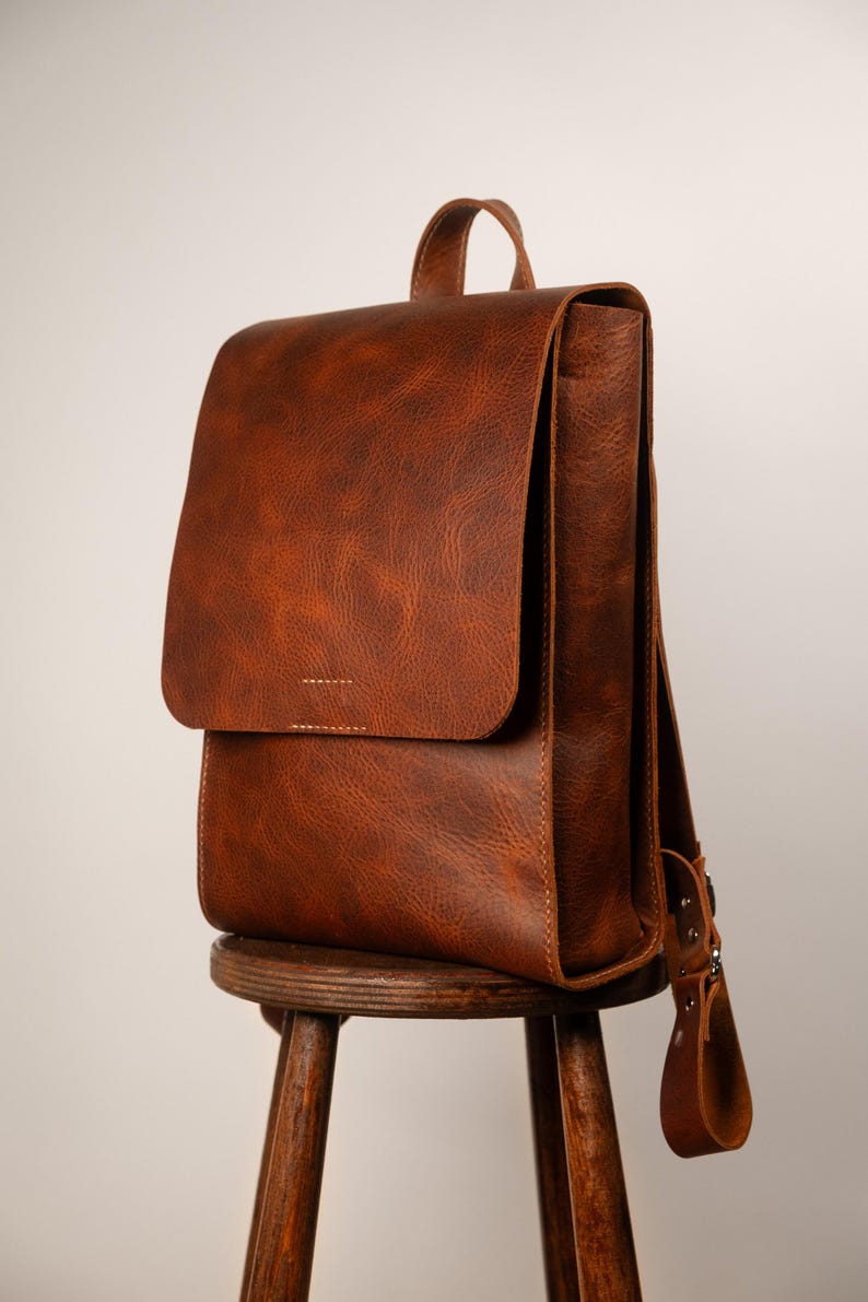 May include: A brown leather backpack with a flap closure, handle, and adjustable straps. The bag is sitting on a wooden stool. The leather has a textured appearance.