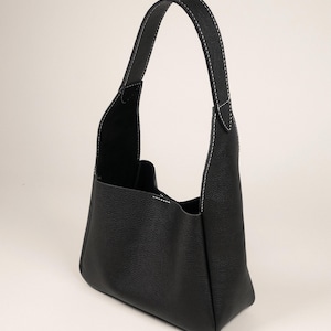 May include: Black leather handbag with a single handle. The bag features white stitching along the seams and handle. The bag has a rectangular base and a spacious interior. The bag is made of a textured leather material.
