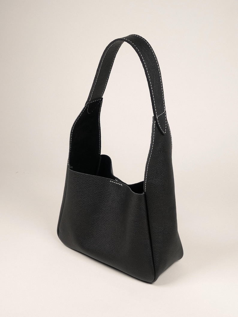 May include: Black leather handbag with a single handle. The bag features white stitching along the seams and handle. The bag has a rectangular base and a spacious interior. The bag is made of a textured leather material.