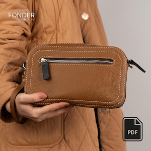 Puede incluir: Un bolso bandolera de cuero marrón con cierre de cremallera y costuras blancas. El bolso tiene un tirador de cremallera negro y una correa de muñeca. El bolso es rectangular con esquinas redondeadas. La marca "FONDER Atelier" es visible.