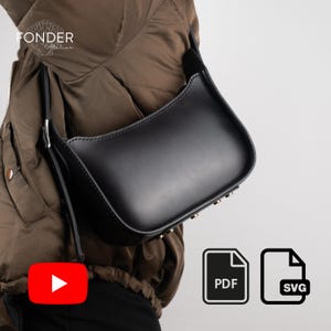 Op de afbeelding: Een zwarte leren crossbody tas met een gebogen ontwerp. De tas heeft een lange, dunne riem en kleine goudkleurige studs aan de onderkant. De tas wordt over een bruine jas gedragen. De afbeelding bevat ook de tekst "FONDER Atelier".