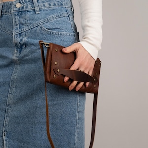 Peut inclure: Un sac bandouli&egrave;re en cuir marron avec une longue sangle et une poign&eacute;e. Le sac est rectangulaire avec des rivets en laiton et une fermeture &agrave; rabat. Le sac est tenu par une personne portant une chemise blanche &agrave; manches longues et une jupe en jean.