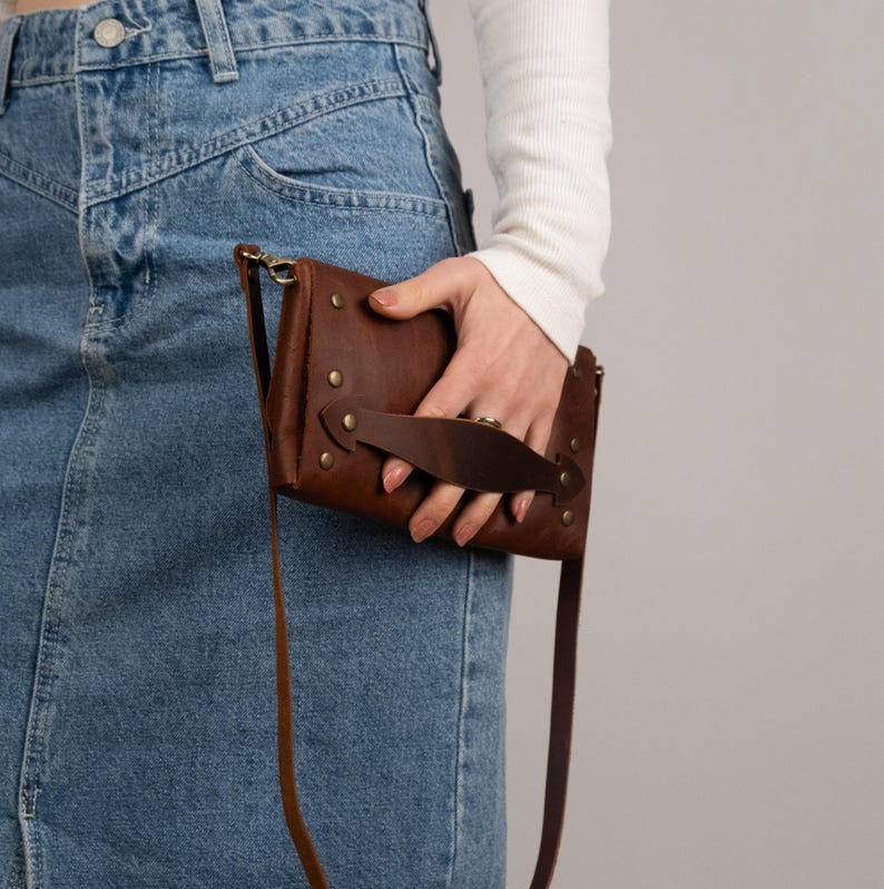 Peut inclure: Un sac bandouli&egrave;re en cuir marron avec une longue sangle et une poign&eacute;e. Le sac est rectangulaire avec des rivets en laiton et une fermeture &agrave; rabat. Le sac est tenu par une personne portant une chemise blanche &agrave; manches longues et une jupe en jean.