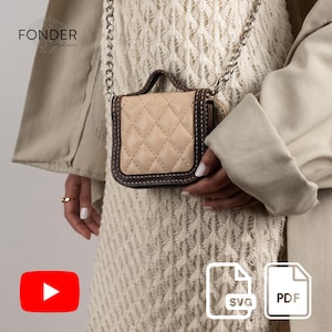 May include: A small, beige quilted handbag with a dark brown trim and a silver chain strap. The bag has a top handle and is held by a person. The bag is a square shape with a diamond pattern. The bag is a neutral color.