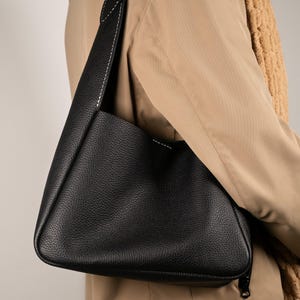 May include: A black leather shoulder bag with a textured surface and a long strap. The strap is stitched with white thread. The bag is worn over a tan coat and a brown scarf.