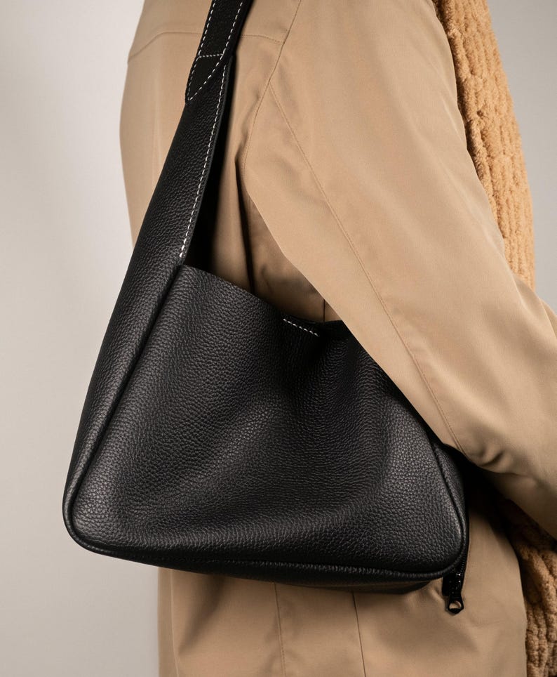 May include: A black leather shoulder bag with a textured surface and a long strap. The strap is stitched with white thread. The bag is worn over a tan coat and a brown scarf.