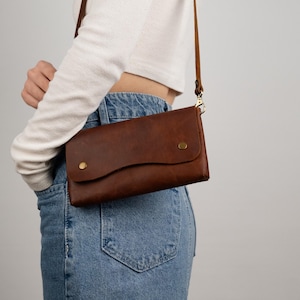 Peut inclure: Un sac bandouli&egrave;re en cuir marron avec une fermeture &agrave; rabat et des accessoires dor&eacute;s. Le sac est dot&eacute; d'une longue bandouli&egrave;re r&eacute;glable et de deux petits rivets dor&eacute;s sur le devant. Le sac est port&eacute; &agrave; l'&eacute;paule.