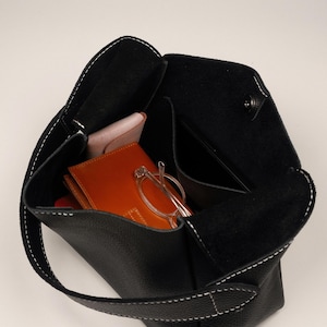 May include: Black leather tote bag with white stitching. The bag is open, revealing a wallet, glasses, and a phone. The bag has a single handle.