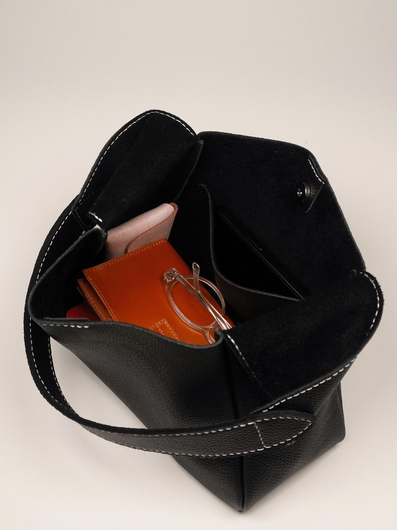 May include: Black leather tote bag with white stitching. The bag is open, revealing a wallet, glasses, and a phone. The bag has a single handle.