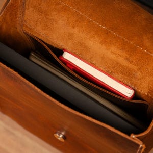 May include: A brown leather satchel with a flap closure, showcasing a spacious interior. Inside, a black laptop, a stack of papers, and a red notebook are visible. The bag has a worn, textured appearance.