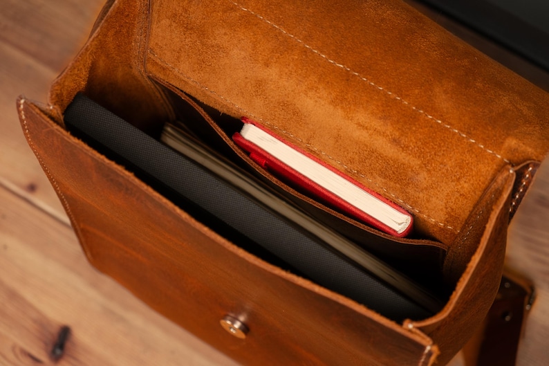 May include: A brown leather satchel with a flap closure, showcasing a spacious interior. Inside, a black laptop, a stack of papers, and a red notebook are visible. The bag has a worn, textured appearance.