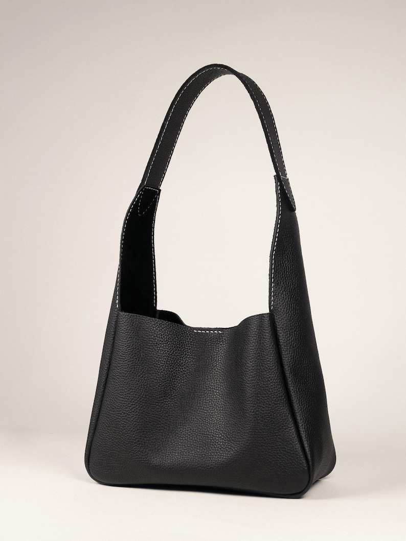 May include: A black leather shoulder bag with a rounded bottom and a single handle. The bag features white stitching along the seams and handle, and has a textured surface.