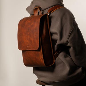 May include: A brown leather backpack with a flap closure and a top handle. The backpack has adjustable shoulder straps and a textured surface. The bag is worn over the shoulder. The person is wearing a gray hoodie.