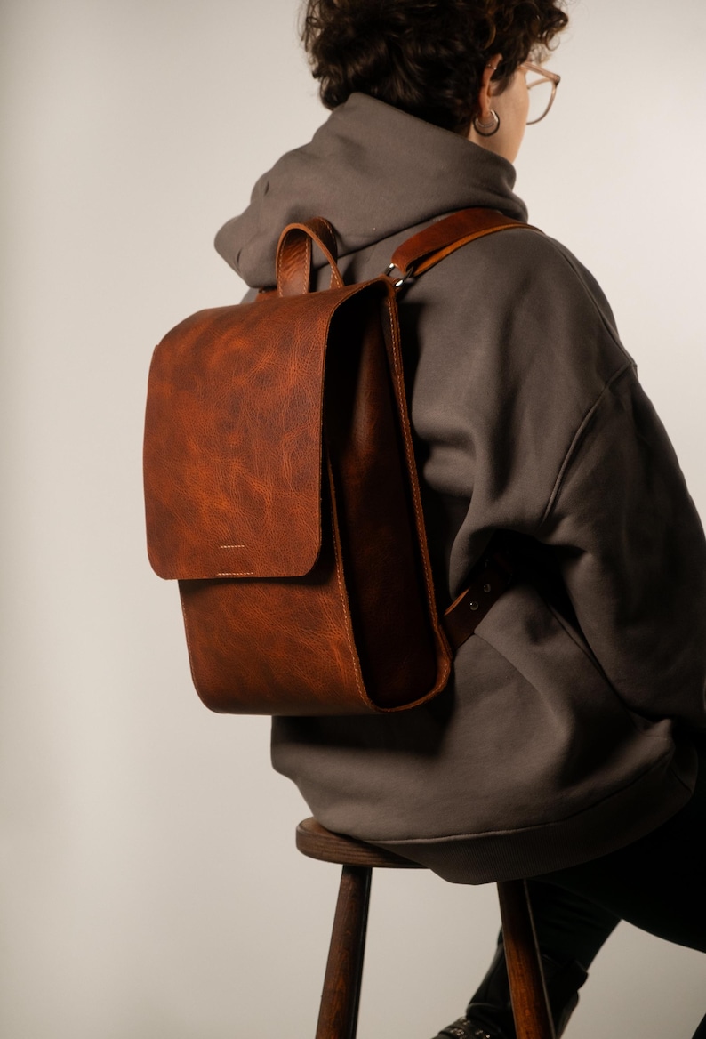 May include: A brown leather backpack with a flap closure and a top handle. The backpack has adjustable shoulder straps and a textured surface. The bag is worn over the shoulder. The person is wearing a gray hoodie.