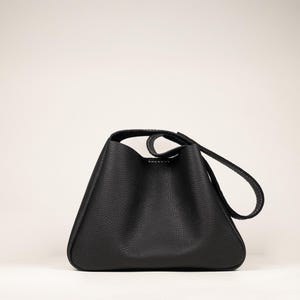 May include: A black leather handbag with a unique triangular shape. The bag features a single handle and a long strap, both with white stitching. The bag has a textured surface and a minimalist design, suitable for everyday use.