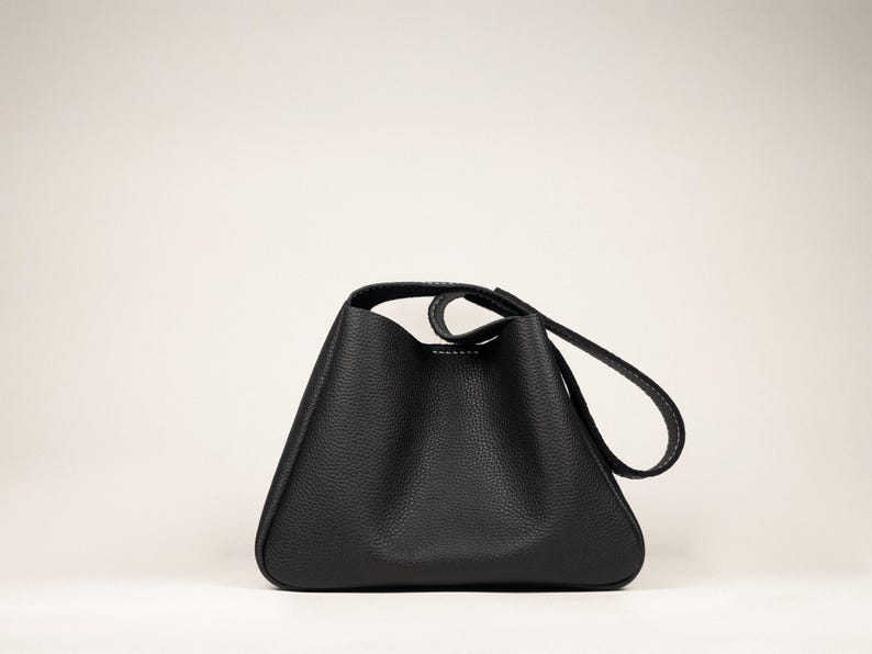 May include: A black leather handbag with a unique triangular shape. The bag features a single handle and a long strap, both with white stitching. The bag has a textured surface and a minimalist design, suitable for everyday use.