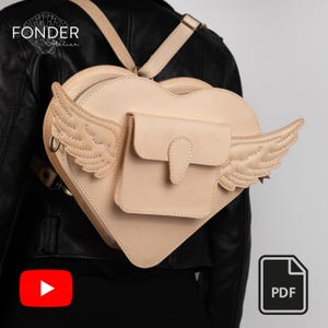 Puede incluir: Mochila en forma de corazón de color beige claro, con detalles de alas. Tiene un pequeño bolsillo frontal y correas ajustables. La palabra "FONDER" es visible en la parte superior. La mochila se lleva sobre una chaqueta de cuero negra.
