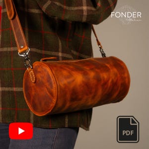 Peut inclure: Sac en cuir marron de forme cylindrique avec une bandoulière. Le sac présente une finition vieillie. Il est présenté sur une chemise à carreaux. Fermeture éclair et accessoires métalliques. Le sac porte le texte "FONDER Atelier".