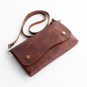 Peut inclure: Un sac bandouli&egrave;re en cuir marron avec une fermeture &agrave; rabat fix&eacute;e par deux clous couleur laiton. Le sac a une longue et fine bandouli&egrave;re et une forme rectangulaire. Le cuir a un aspect textur&eacute;.