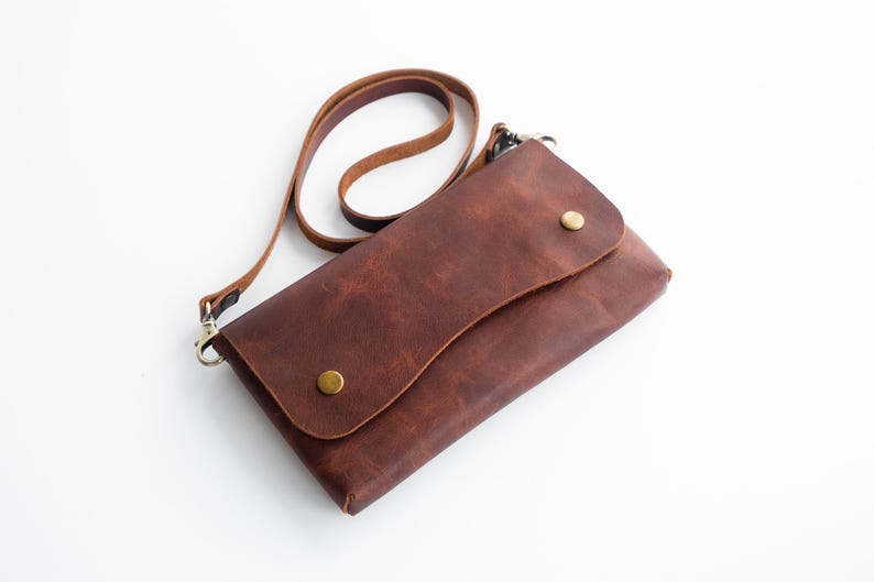 Peut inclure: Un sac bandouli&egrave;re en cuir marron avec une fermeture &agrave; rabat fix&eacute;e par deux clous couleur laiton. Le sac a une longue et fine bandouli&egrave;re et une forme rectangulaire. Le cuir a un aspect textur&eacute;.