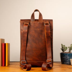 May include: A brown leather backpack with adjustable shoulder straps and a top handle. The bag has a rectangular shape with a flap closure and silver-colored metal hardware. Books and a potted cactus are visible in the background.