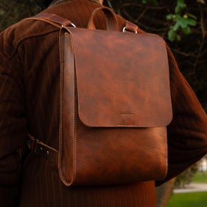 May include: A brown leather backpack with a flap closure and adjustable straps. The bag has a rectangular shape with a top handle and is worn on the back. The leather has a textured appearance and is stitched with white thread.