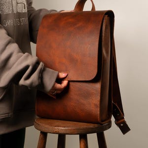 May include: A brown leather backpack with a flap closure and adjustable shoulder straps. The bag has a top handle and is sitting on a wooden stool. The leather has a textured appearance.