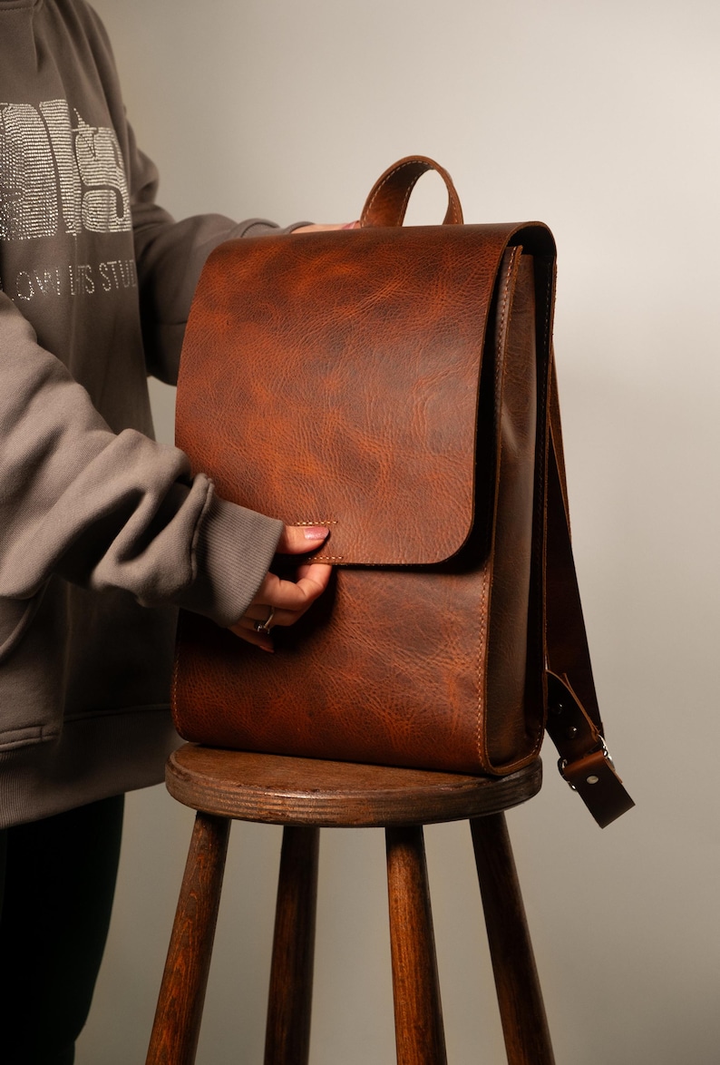 May include: A brown leather backpack with a flap closure and adjustable shoulder straps. The bag has a top handle and is sitting on a wooden stool. The leather has a textured appearance.