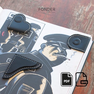 Op de afbeelding: Zwarte leren hoekboekmarkeringen met witte stiksels op een boek met een stripillustratie. Eén bladwijzer is hartvormig, een andere rechthoekig en de derde een afgeronde rechthoek. Het woord "FONDER" staat bovenaan.