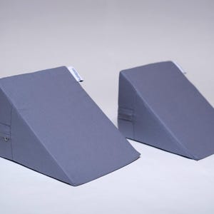 May include: Two gray wedge pillows are displayed against a white background. The pillows are triangular, with a sloped top and a flat bottom. Each pillow has a small white tag on the side. The pillows are made of a soft, textured fabric.