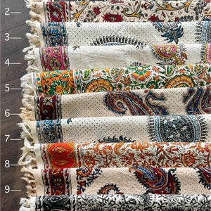 May include: A stack of ten decorative fabric runners with fringed edges. Each runner features a unique pattern in various colors, including paisley, floral, and geometric designs. The color palette includes shades of orange, blue, red, and black.