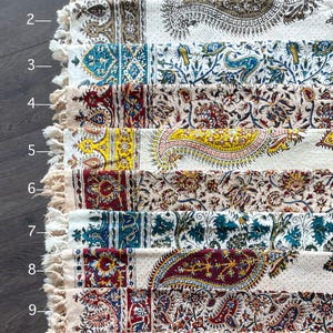 May include: A collection of ten rectangular, patterned textiles with fringed edges. Each textile features a unique design with paisley and floral motifs in various colors, including red, blue, yellow, and brown, set against a cream background. The textiles are arranged side by side.