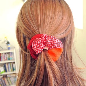 Puede incluir: Un accesorio para el cabello rojo y naranja, probablemente una goma, está atado en el cabello. El accesorio presenta un diseño estampado con cuadrados blancos sobre un fondo rojo y una sección naranja sólida.