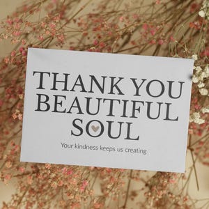 Könnte beinhalten: Eine weiße Karte mit den Worten "THANK YOU BEAUTIFUL SOUL" in schwarzem Text, mit einem Herzsymbol. Der Text "Your kindness keeps us creating" steht darunter. Die Karte ist von getrockneten Blumen umgeben.