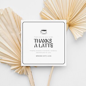 Könnte beinhalten: Eine quadratische weiße Karte mit schwarzem Text, der "THANKS A LATTE" und "BREWED WITH LOVE" lautet. Die Karte ist von zwei getrockneten Palmblättern umgeben, was eine neutrale und minimalistische Ästhetik schafft.