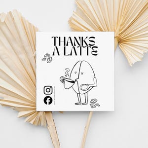 Könnte beinhalten: Eine quadratische weiße Karte mit dem Text "THANKS A LATTE" und einer Cartoon-Kaffeebohnenfigur, die eine Tasse Kaffee hält. Die Karte enthält auch Instagram- und Facebook-Symbole. Der Hintergrund umfasst getrocknete Palmblätter.