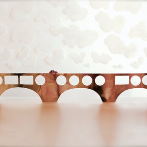 Puede incluir: Un coche rojo conduce a través de un puente marrón con círculos y cuadrados blancos. El puente está sobre un fondo blanco con nubes blancas.