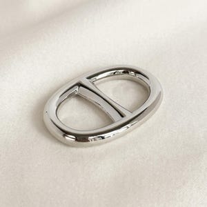 May include: A polished silver-tone metal buckle featuring an oval frame with a central horizontal bar. The hardware has a smooth, reflective surface and a rounded, continuous design, showcasing a clean and simple aesthetic for various fastening needs.
