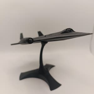 Puede incluir: Un modelo negro de un avión SR-71 Blackbird, montado sobre un soporte curvo. El modelo presenta alas detalladas, aletas de cola y una cabina. El soporte tiene una base decorativa. El modelo de avión mide aproximadamente 30 cm de largo.