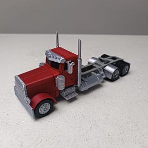 May include: A red and silver toy articulated lorry with a detailed design. The lorry features a red cab with silver accents on the grill, fenders, and roof. The trailer hitch and wheels are silver. It has two exhaust pipes and a fuel tank.