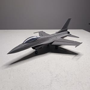 May include: A gray model airplane with a sleek design, featuring a pointed nose, wings, and a tail fin. The toy aircraft has a clear cockpit and is resting on a white surface. The model is a detailed replica of a fighter jet.