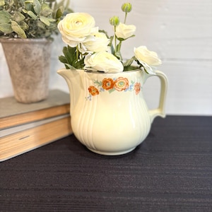 Może przedstawiać: Kremowy ceramiczny dzbanek z kwiatowym wzorem w kolorze pomarańczowym i niebieskim. Dzbanek jest wypełniony białymi kwiatami. Dzbanek ma uchwyt i dzióbek. Dzbanek stoi na ciemnej powierzchni.