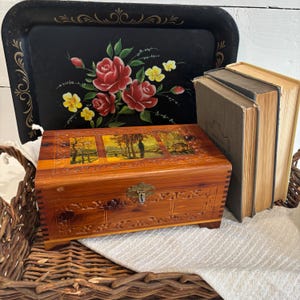 May include: A black serving tray with a floral design featuring red roses and yellow flowers. A wooden cedar chest with a landscape painting on the lid and a metal clasp. Two vintage books are next to the chest.